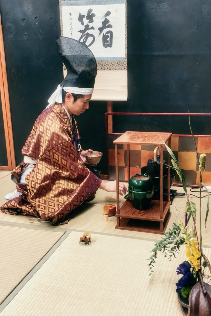 Chris Yao in hiratare and eboshi preparing tea beside a wooden shelf, tea utensils, and flowers at Ogasawara-ryū America